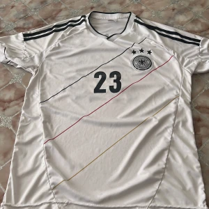 Tysklands fotbollströja Gomez #23 - Vit Tyskland fotbollströja från Adidas med svarta detaljer, nummer 23 och namnet Gomez på ryggen. Tröjan har korta ärmar, broderat landslagsmärke och tre stjärnor ovanför loggan. Tunt och sportigt material, perfekt för match eller träning.