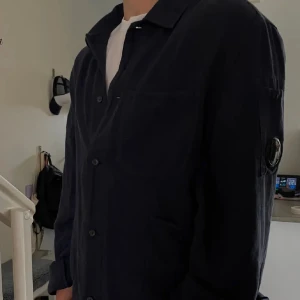 Mörkblå overshirt från C.P. Company - Säljer en mörkblå overshirt från C.P. Company med klassisk krage, knäppning framtill och flera fickor. Ikonisk linsdetalj på ärmen ger jackan en unik vibe. Tillverkad i bomull och har lång ärm. Perfekt för lager-på-lager-stil. Passar både M och L!