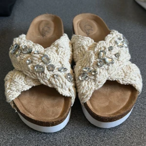 Beige sandaler med flätad ovandel och stenar - Snygga sandaler från Clarks med flätad beige ovandel i stickad stil och stora glittrande stenar. De har en plattformsula i vitt och brunt som ger extra höjd och en öppen tå för sommarens alla outfits.