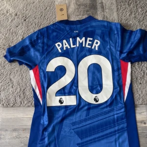 Chelsea Palmer 20 matchtröja Nike. Den är helt ny Chelsea tröja till säsongen 25/26. . - Chelsea FC hemmatröja med Palmer 20-tryck. Klassisk blå färg med vita detaljer och diskreta ränder, officiellt klubbmärke på bröstet och Nike-logga. Tillverkad i lätt och ventilerande polyester, perfekt för match eller supporterliv. Mycket bra skick. Har ej använts innan!. 