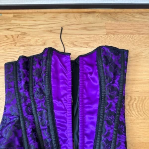 Lila korsett från Burleska med spets - Säljer en lila och svart korsett från Burleska med snygga svarta spetsdetaljer och snörning i ryggen. Satinliknande material med blommigt spetsmönster och markerade svarta sömmar. Perfekt för dig som vill sticka ut med en edgy och cool look.