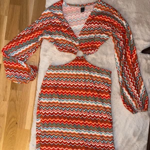 Färgglad zigzagklänning med cut out - Säljer en kort klänning från Bershka med färgglatt zigzag-mönster i rött, orange, blått och vitt. Klänningen har lång ärm, v-ringning och en snygg cut out med ringdetalj framtill. Perfekt för dig som vill sticka ut med en unik look!