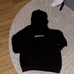 Svart hoodie från Seams med tryck - Svart hoodie från Seams i storlek M med vit broderad logga på bröstet och liten stjärndetalj på ärmen. Klassisk passform med huva och känguruficka. Mjuk insida och stilren look, perfekt för dig som gillar streetwear.
