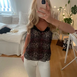 Leopardmönstrad linnetopp med spets - Snygg och luftig linnetopp med leopardmönster i svart, rött och beige. Toppen har tunna axelband och svart spets längs urringningen för en extra detalj. Perfekt att styla med jeans eller kjol för en trendig look.