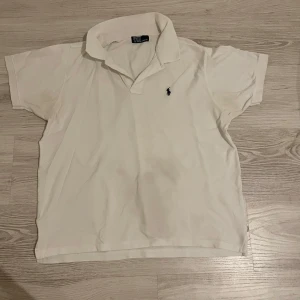 Vit pikétröja från Polo Ralph Lauren - Klassisk vit pikétröja från Polo Ralph Lauren med korta ärmar och broderad marinblå logga på bröstet. Tröjan har krage och knäppning framtill. Perfekt för en clean och stilren look.