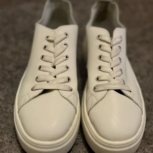Vita sneakers med platt sula - Stilrena vita sneakers i skinn med rund tå och platt sula. Skorna har klassisk snörning med vita skosnören och silverfärgade öljetter. Perfekta för en clean och modern look, passar till det mesta i garderoben.