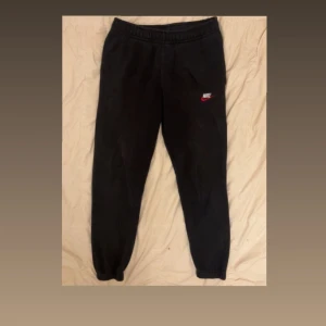 Svarta mjukisbyxor från Nike - Svarta mjukisbyxor från Nike med broderad logga i vitt och rött på vänster ben. Byxorna har elastisk midja med snörning och muddar vid bensluten. Mjukt material och en bakficka. Perfekta för chill eller träning.