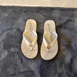 Vita flip-flops från Michael Kors - Snygga vita flip-flops från Michael Kors med guldigt MK-emblem på remmen. Sulan har ett diskret mönster och signaturtryck. Perfekta för sommardagar och strandhäng, med klassisk och stilren design.