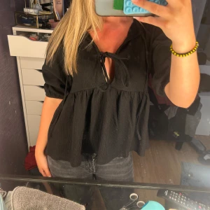 Svart blus med knyt och puffärm - Säljer en svart blus med snygg knytning framtill och lätt v-ringning. Toppen har puffiga trekvartsärmar och ett flowy, avslappnat fall. Perfekt för dig som gillar en trendig och chill look.