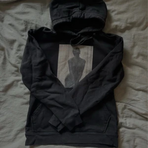 Svart hoodie med konstmotiv - Svart hoodie med huva och känguruficka framtill. På bröstet finns ett tryck med konstnärligt motiv av en siluett. Tröjan har långa ärmar och en avslappnad passform, perfekt för en cool och stilren look.