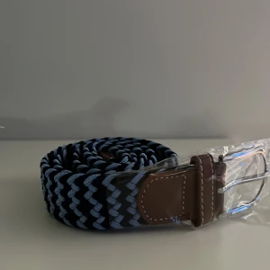 Mörkblått/Ljusblått Flätat bälte - Snyggt flätat bälte i svart och ljusblått med bruna läderdetaljer och silverspänne. Bältet har ett randigt mönster och är perfekt för att ge outfiten en cool touch. Passar till jeans eller chinos och har en flexibel, vävd design.