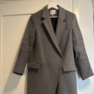 Grå ullkappa från Dagmar, stl 34 - Nypris 4999kr Snygg grå kappa från Dagmar i klassisk rak modell med bred krage och två fickor framtill. Tillverkad i ullblandning. Stilren och tidlös design som passar till många outfits.  