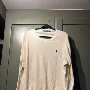 Vit v-ringad tröja Polo Ralph Lauren - Vit v-ringad tröja från Polo Ralph Lauren i mjuk pima bomull. Klassisk design med broderad logga på bröstet och långa ärmar. Perfekt för en clean och stilren look, passar till jeans eller chinos.