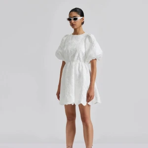 Vit kortklänning By Malina - Superfin vit kortklänning från By Malina med puffiga kortärmar, broderade blommor och öppen rygg med vågig kant. Klänningen har markerad midja och rund halsringning. Perfekt för studenten! Endast använd 1 gång, nypris 2800kr