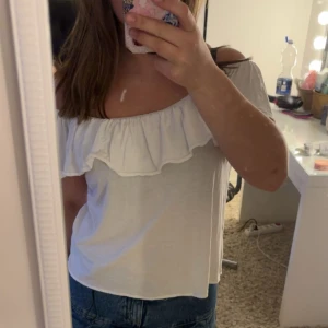 Vit offshoulder topp med volang - Säljer en söt vit offshoulder topp med bred volang runt hela överdelen. Toppen är ärmlös och har en luftig passform, perfekt för varma dagar. Materialet känns mjukt och lätt mot huden. Superfin att matcha med jeans eller kjol!