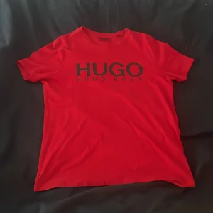 Röd t-shirt från Hugo Boss - Snygg röd t-shirt från Hugo Boss med stor svart logga framtill. Klassisk rund halsringning och korta ärmar. Perfekt för dig som gillar stilrena och trendiga plagg med tydlig branding. Mjuk och skön känsla, passar till det mesta.