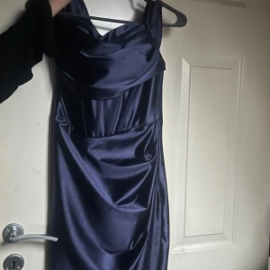 Mörkblå långklänning i satin - Säljer en elegant mörkblå långklänning i glansig satin med draperad urringning och breda axelband. Klänningen har en figurnära passform och ett stilrent, tidlöst snitt som ger en lyxig känsla. Perfekt för dig som vill sticka ut med en klassisk look.