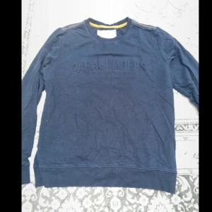 Mörkblå sweatshirt från Parajumpers XL - Mörkblå sweatshirt från Parajumpers i storlek XL. Tröjan har logga på bröstet och ett snyggt PJS-märke på ena ärmen. Klassisk rund halsringning och långa ärmar. Perfekt för dig som gillar stilrena och sportiga plagg.