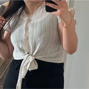 Vit knytblus med knappar - Säljer en vit blus i bomull med korta ärmar och v-ringning. Blusen har knappar framtill och en snygg knytning i midjan. Tyget är lätt strukturerat och ger en avslappnad vibe, perfekt till jeans eller kjol.