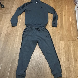 Mörkgrå Adidas hoodie med huva - Mörkgrå hoodie från Adidas med klassiska svarta ränder längs ärmarna och Adidas-logga på bröstet. Tröjan har huva och är långärmad, tillverkad i mjuk bomullsblandning. Perfekt för chill eller träning, snygg och enkel att matcha.