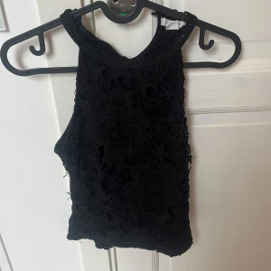 Svart spetsig linnetopp från Amisu - Svart linnetopp från Amisu i storlek S med snyggt spetsmönster framtill. Toppen är ärmlös och har en hög halsringning, vilket ger en cool och trendig look. Perfekt att styla med jeans eller kjol för en extra touch.