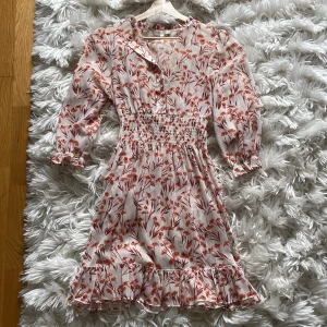 Blommig kortklänning från Maje - Supersöt kortklänning från Maje i vitt med röda och orange blommor med underklänning till. Klänningen har smock i midjan, volangkant nertill, knappar framtill och långa ärmar med rynkade muddar. Perfekt för vår och sommar, i ett lätt och luftigt material.