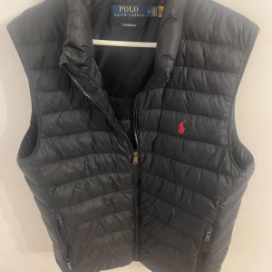 Svart dunväst från Polo Ralph Lauren - Snygg svart dunväst från Polo Ralph Lauren med klassisk röd logga på bröstet. Västen är quiltad, har dragkedja framtill och två fickor med dragkedja. Perfekt för lager-på-lager och funkar till många olika outfits.