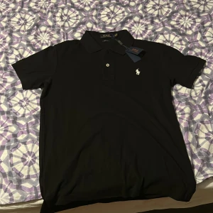 Svart pikétröja från Polo Ralph Lauren - Klassisk svart pikétröja från Polo Ralph Lauren med vit broderad logga på bröstet. Tröjan har korta ärmar, krage och knäppning med två knappar. Tillverkad i mjuk bomull som känns skön mot huden. Perfekt för en clean och stilren look.