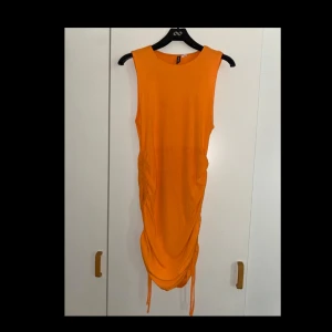 Orange ärmlös klänning  - Snygg orange ärmlös klänning från Divided i storlek L. Modellen är kort med dragsko och rynkade detaljer på sidorna.