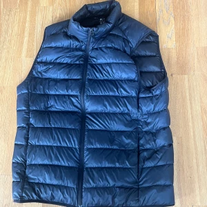 Mörkblå dunväst med dragkedja - Snygg mörkblå dunväst från Uniqlo med quiltad design och hög krage. Västen har en hel dragkedja framtill och är perfekt att slänga över en hoodie eller tröja när det är kyligt ute. Enkel och stilren look som funkar till det mesta.