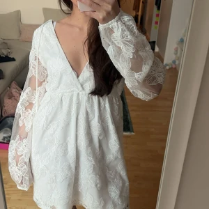 Vit kort spetsklänning med v-ringning, studentklänning - Superfin vit kort klänning med v-ringning och långa transparenta ärmar. Klänningen har broderade blommor i spets över hela tyget och en luftig passform. Perfekt för sommarens alla tillfällen och även för studenten. Den är helt ny!!