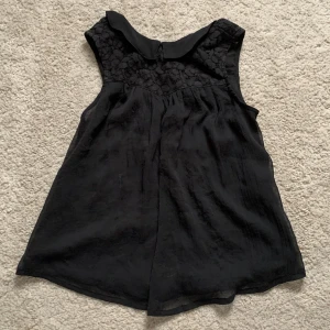 Svart ärmlös blus med spetsdetalj - Supersöt svart blus i luftig modell med ärmlös design. Ovanför bysten finns en snygg spetsdetalj som ger en extra touch. Blusen har en liten krage och lätt utställd passform, perfekt för dig som gillar stilrena och feminina plagg.👏🏻