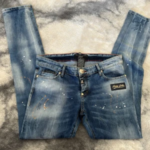 Philipp Plein skinny jeans med stänk - Snygga blå skinny jeans från Philipp Plein med coolt stänkmönster i vitt och orange. Jeansen har klassisk femficksdesign, guldiga detaljer och logopatch både fram och bak. Perfekt för dig som vill sticka ut med en unik streetstyle.