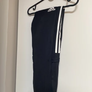 Svarta Adidas träningsbyxor med ränder - Svarta träningsbyxor från Adidas med klassiska vita ränder längs sidorna och Adidas-logga på låret. Byxorna har elastisk midja och är gjorda i ett syntetmaterial som passar perfekt för sport och träning. Snygg och sportig look som funkar till allt. Passar runt 165-170 