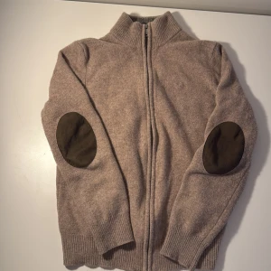 Beige kofta i lammull från Hackett - Snygg beige kofta från Hackett i 100% lammull med hel dragkedja och ribbad krage. Mörkare förstärkningar på armbågarna ger en cool touch. Perfekt för lager-på-lager och chill höst- eller vinterstil. Står m men är xs passar även s