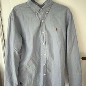 Ljusblå skjorta Polo Ralph Lauren L - Klassisk ljusblå skjorta från Polo Ralph Lauren i slim fit-modell. Skjortan har button down-krage, broderad logga på bröstet och vita knappar hela vägen. Tillverkad i bomull och har långa ärmar. Perfekt för en clean och stilren look.