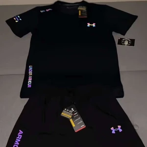 Svart t-shirt och shorts från Under Armour - Svart t-shirt och shorts från Under Armour med kort ärm och färgglada loggor på bröstet och ärmen. Snyggt tryck med märkesnamnet längs sidan. Perfekt för dig som gillar sportig och clean stil. Materialet känns lätt och andas bra. Helt oanvänd