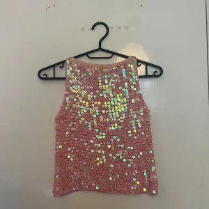 Rosa paljettopp från Lipsy - Supersöt rosa topp från Lipsy, täckt med skimrande fyrkantiga paljetter som fångar ljuset. Stickad bas i rosa med ärmlös design och rund hals. Perfekt för dig som vill sticka ut och glänsa lite extra.