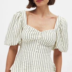 Randig blus med puffärm och rosett - Superfin blus i ljusbeige med svarta ränder, puffiga korta ärmar och söt knytrosett i ryggen. Toppen har markerad midja, knappar framtill och fyrkantig ringning. Perfekt för dig som gillar romantisk och trendig stil. Passar till s-m🩷