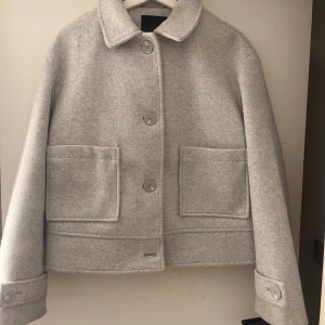Grå ulljacka med stora fickor - Snygg grå ulljacka med stora knappar och två rejäla fickor framtill. Jackan har en klassisk krage och långa ärmar med knappdetaljer vid ärmsluten. Perfekt för vårväder. Köpare står för frakt.