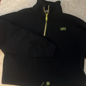 Svart fleece tröja från Levi's - Säljer en svart fleece tröja från Levi's med halv dragkedja i gult och justerbar dragsko i nederkant. Perfekt för kyliga dagar med sin mjuka och varma känsla. Levi's logga i gult på bröstet ger en snygg kontrast.