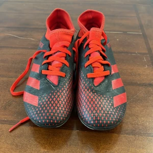 Adidas Predator fotbollsskor - Snygga Adidas Predator fotbollsskor i svart och rött med klassiska ränder och snörning. Perfekta för fotbollsplanen med en tight passform och greppvänlig sula.