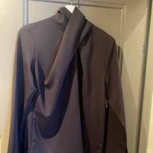 Grå långärmad blus med draperad krage - Mörkgrå blus från H&M i storlek S. Hög, asymmetriskt draperad hals och långa trumpetärmar med slit. Knäpps med tre dolda knappar i nacken (bild 5). Nyskick. Färgen är mörkgrå och slår inte åt lila som det ser ut på bilderna.