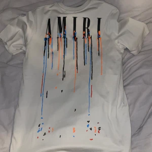 Beige t-shirt från Amiri med  - Säljer en Beige t-shirt från Amiri med coola färgstänk i blått, orange och svart. T-shirten har korta ärmar och ett tryck med Amiri-loggan både fram och bak. Perfekt för en avslappnad stil! Har aldrig använts!
