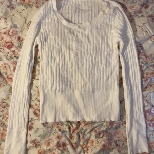 Vit ribbad tröja med knappar - Säljer en stilren vit ribbad tröja med knappar framtill. Tröjan har långa ärmar och är tillverkad i en mjuk polyesterblandning. Perfekt för en enkel och snygg look.
