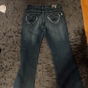 Blå low waist bootcut jeans med tryck på fickorna  - Snygga blå jeans från Miss Me med unika detaljer på bakfickorna i form av hjärtan och broderier. Jeansen har en klassisk femficksdesign och en bekväm passform. Perfekta för dig som vill ha något extra i garderoben!