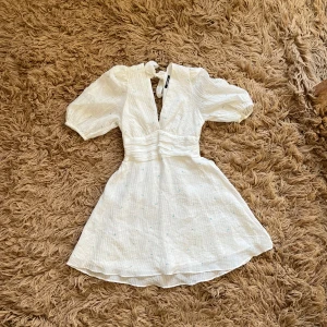 Vit kortklänning med puffärmar - Vit kortklänning med puffiga kortärmar, knytning i nacken och så blåa blommor på. Nyskick, använd 1 gång.