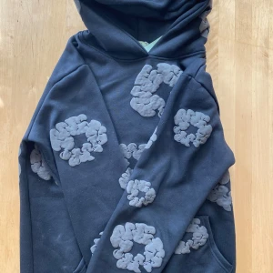 Mörkgrå hoodie från Denim Tears - Säljer en mörkgrå hoodie från Denim Tears med unikt mönster i ljusare grått. Hoodien har en klassisk design med känguruficka och en bekväm huva. Perfekt för en avslappnad stil. Priset är ej hugget