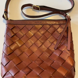 Brun axelväska i buffelläder från Saddler - Brun axelväska i buffelläder från Saddler med flätad design. Väskan har en justerbar axelrem och dragkedja upptill. Insidan är fodrad med polyester och har två  innerfack, en med dragkedja.