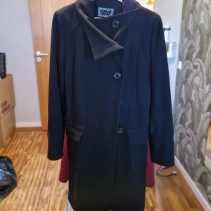 Svart kappa med knappar - Elegant svart kappa med knappar framtill och ett bälte i midjan. Den har långa ärmar och en klassisk krage. Perfekt för kyligare dagar och ger en stilren look.